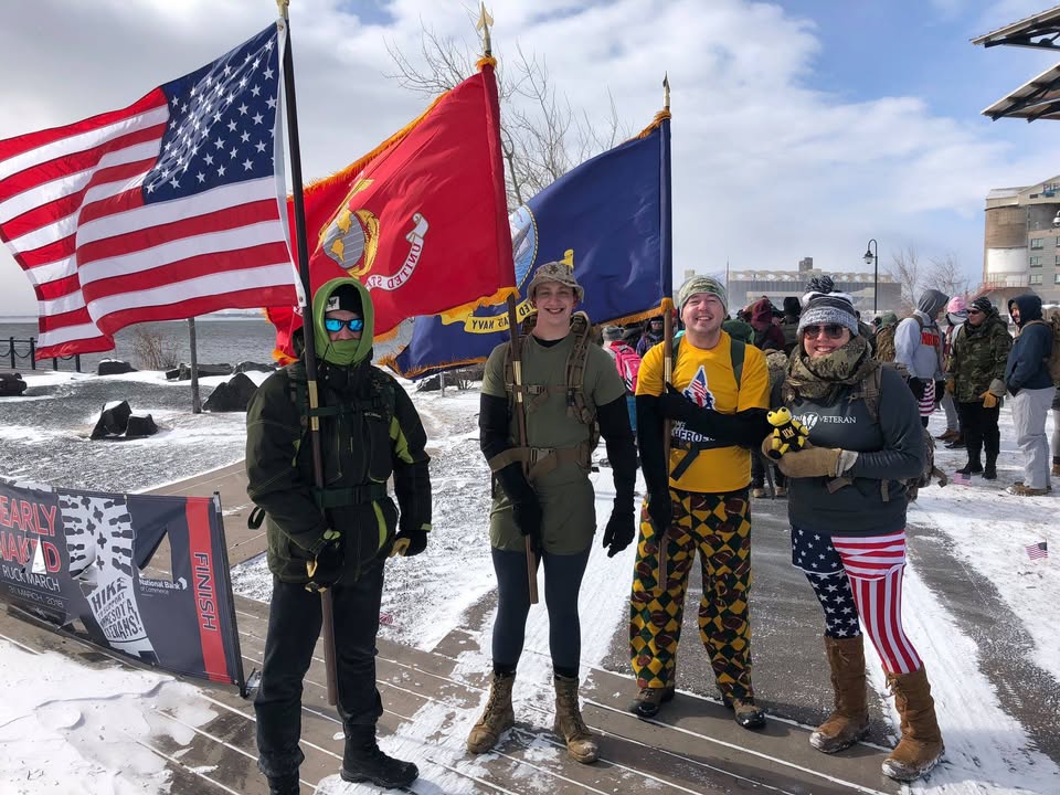 Four people stand outdoors in the snow holding American, Marine Corps, and military branch flags at the finish line of a ruck march.