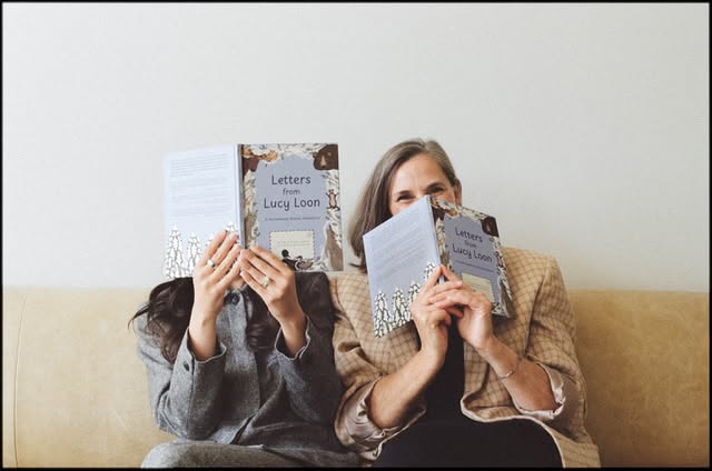 Two women sitting on a sofa, each holding a copy of the book “Letters from Lucy Loon” in front of their faces.